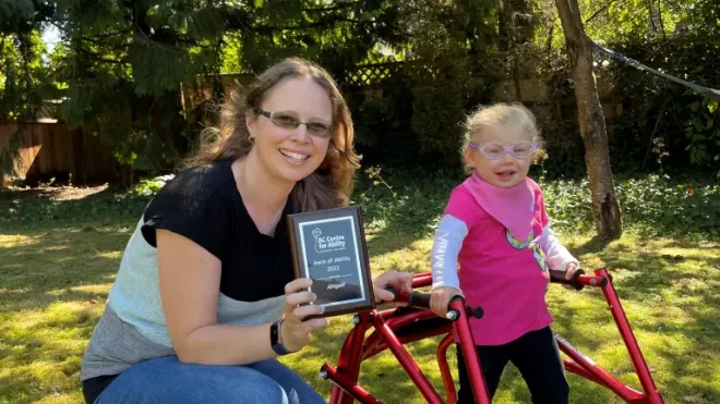 Abby outside using her walker with her Mom holding her Hero of Ability Award 