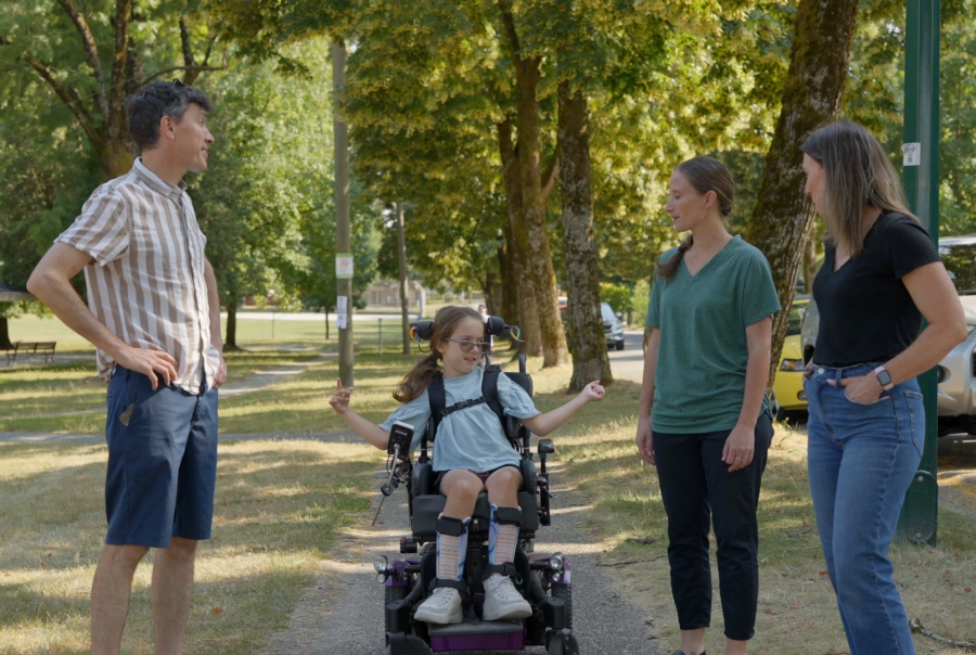 Three adults and a child in a wheelchair conversing in a park.